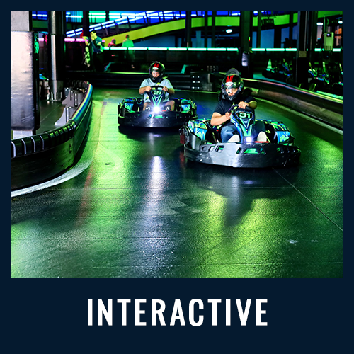 Two people racing go-karts on an indoor neon-lit track with "INTERACTIVE" text below in white