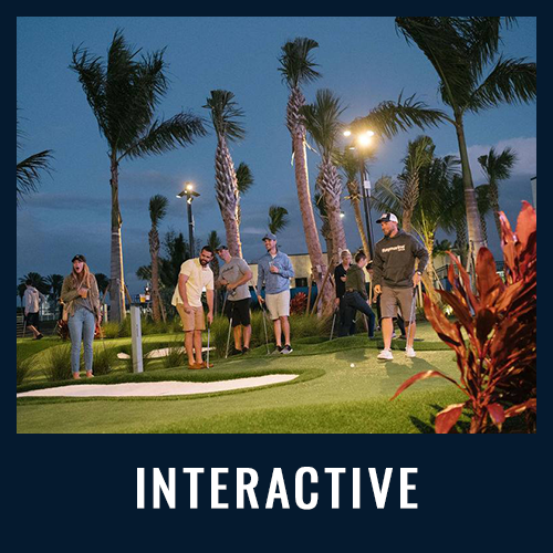 People playing mini-golf at night on a tropical-themed course with palm trees, labeled “INTERACTIVE”