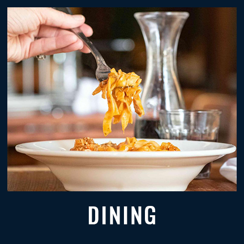 A hand twirls pasta with a fork over a bowl of pasta and meat sauce, with a glass and wine
