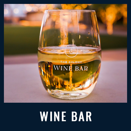 Glass of white wine with "The Colony Wine Bar" logo, set on a table with soft evening lights