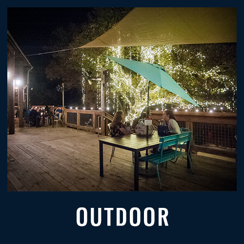 People dining on a wooden patio at night under string-lit trees and shade sails