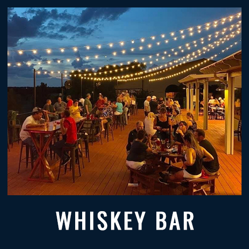 People enjoy drinks and conversation under string lights at an outdoor whiskey bar during sunset
