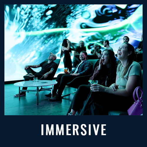 Guests watch an immersive digital light show in a dark room, seated in lounge chairs