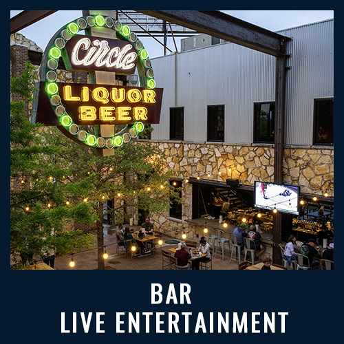 Outdoor bar with a neon sign, string lights, people dining and a large screen showing sports