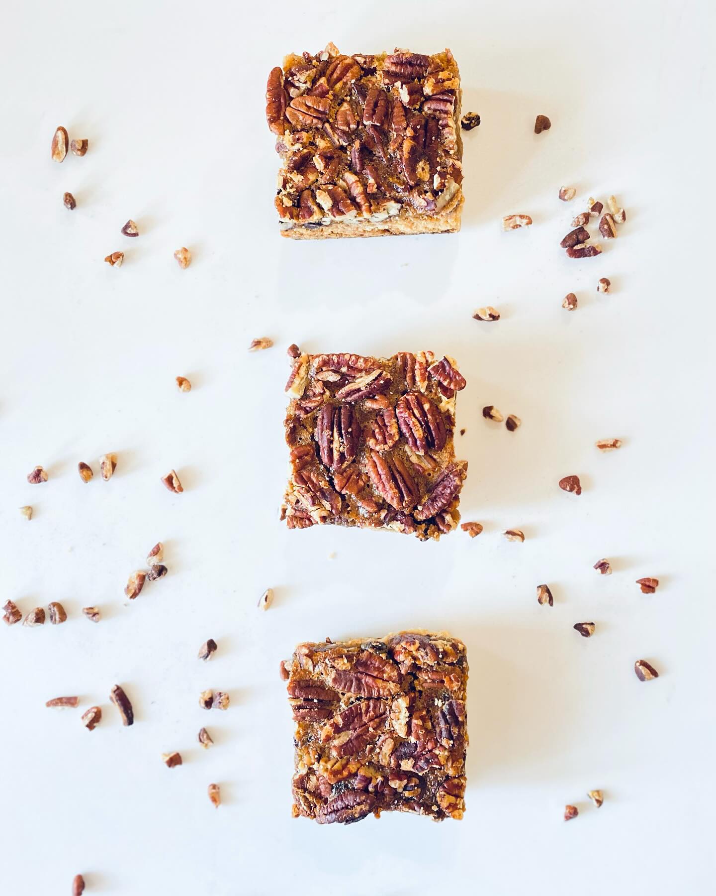 Three pecan pie bars arranged vertically on a white surface with scattered pecan pieces.