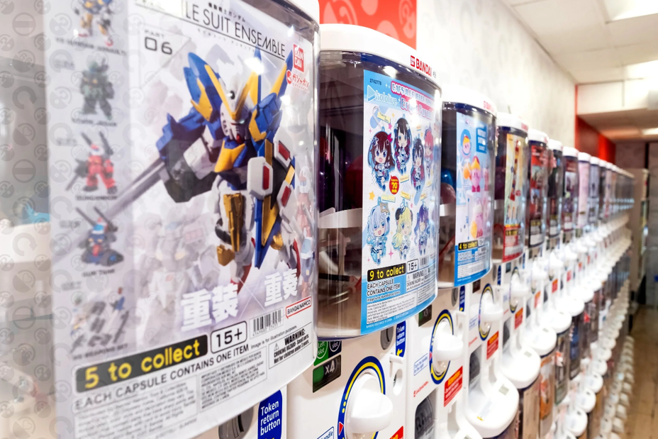 Row of Japanese capsule toy machines (Gashapon) featuring collectible anime and robot figures.